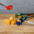 Colorful silicone tongs with hand-shaped grips picking up cheese cubes on a wooden board.