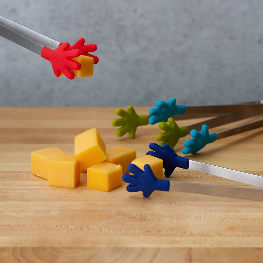 Colorful silicone tongs with hand-shaped grips picking up cheese cubes on a wooden board.