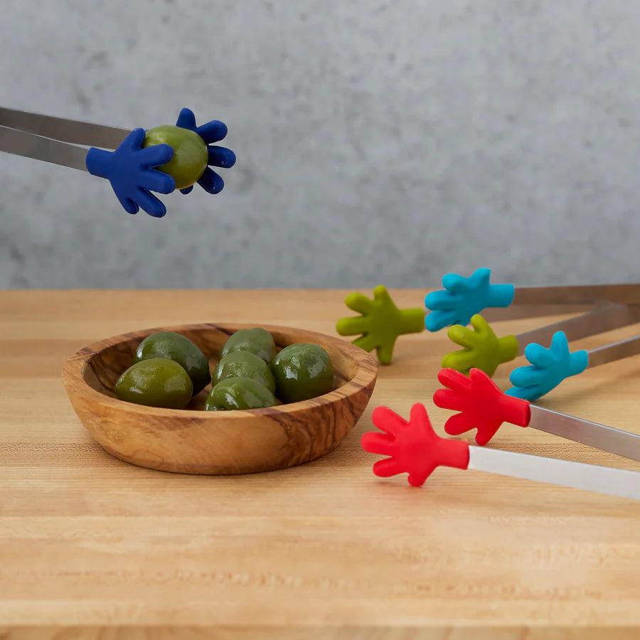 Colorful tongs with hand-shaped ends picking up olives from a wooden bowl on a wooden surface.
