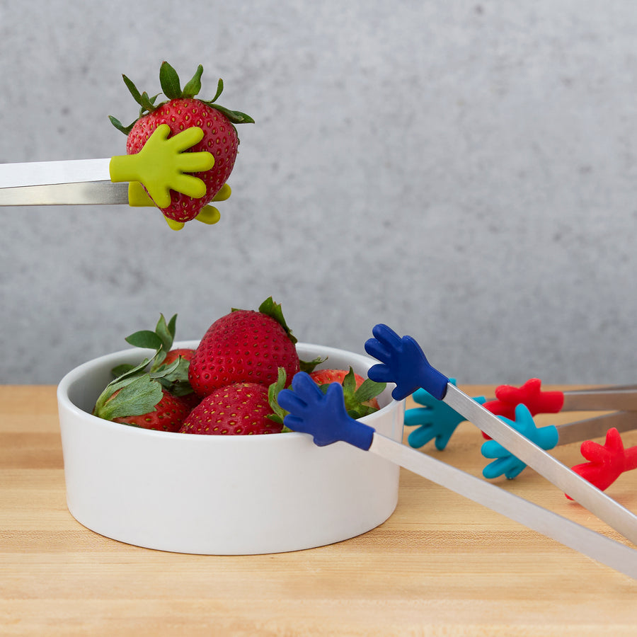 Tongs with hand-shaped grips on a wood surface with a bowl of strawberries and one air of tongs holding a strawberry.