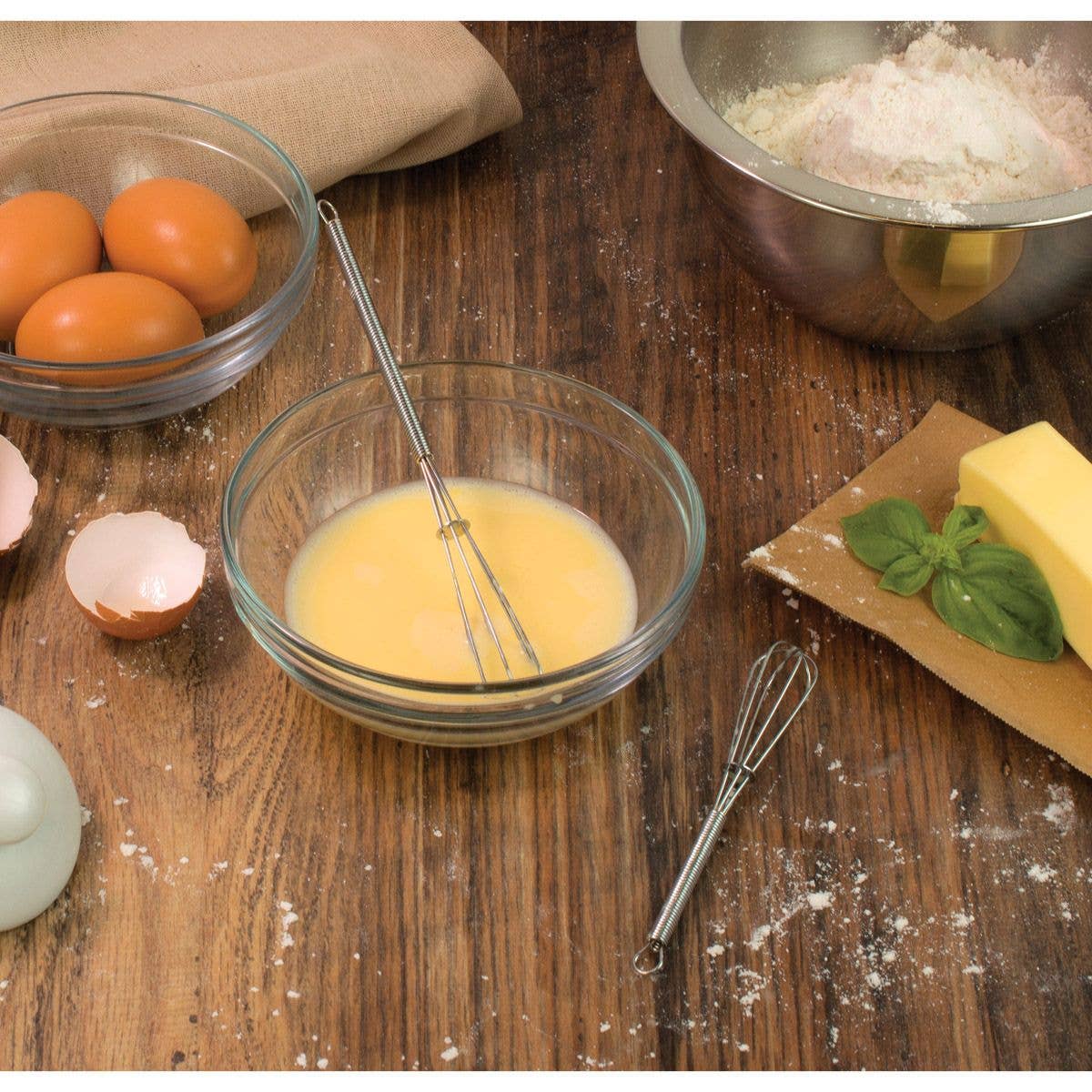 Bowl of whisked eggs with a whisk on a wooden table, surrounded by ingredients.