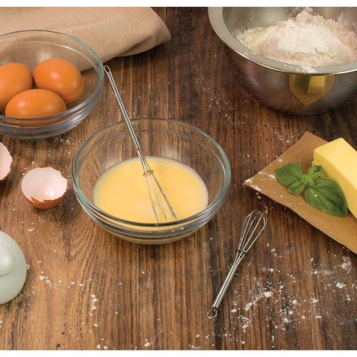 Bowl of whisked eggs with a whisk on a wooden table, surrounded by ingredients.
