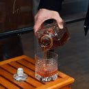 A person pouring liquor into a drinking glass with a geometric texture pattern on a wooden table.