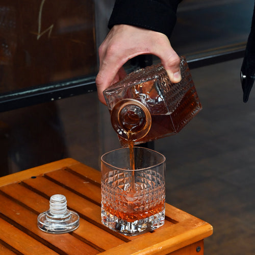 A person pouring liquor into a drinking glass with a geometric texture pattern on a wooden table.