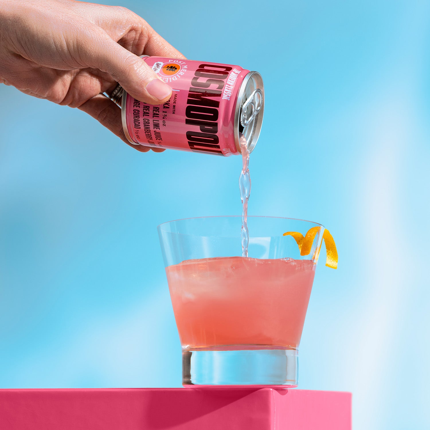 A hand pours a pink cocktail from a pink can labeled 'Cosmopolitan' into a clear glass filled with pink drink and an orange peel garnish, set against a bright blue background with a pink pedestal.
