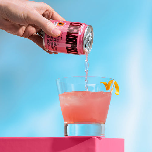 A hand pours a pink cocktail from a pink can labeled 'Cosmopolitan' into a clear glass filled with pink drink and an orange peel garnish, set against a bright blue background with a pink pedestal.