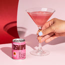 A hand holds a pink cocktail in a martini glass next to a pink can of Cosmopolitan drink on a pink and red background.