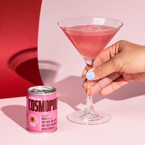 A hand holds a pink cocktail in a martini glass next to a pink can of Cosmopolitan drink on a pink and red background.