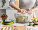 A person is using a transparent salad spinner with a non-slip base. The spinner is filled with salad, and there are other kitchen tools and a bowl with more salad ingredients in the background.