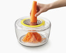 Hand using a carrot grater to shred a carrot into a clear container on a white background