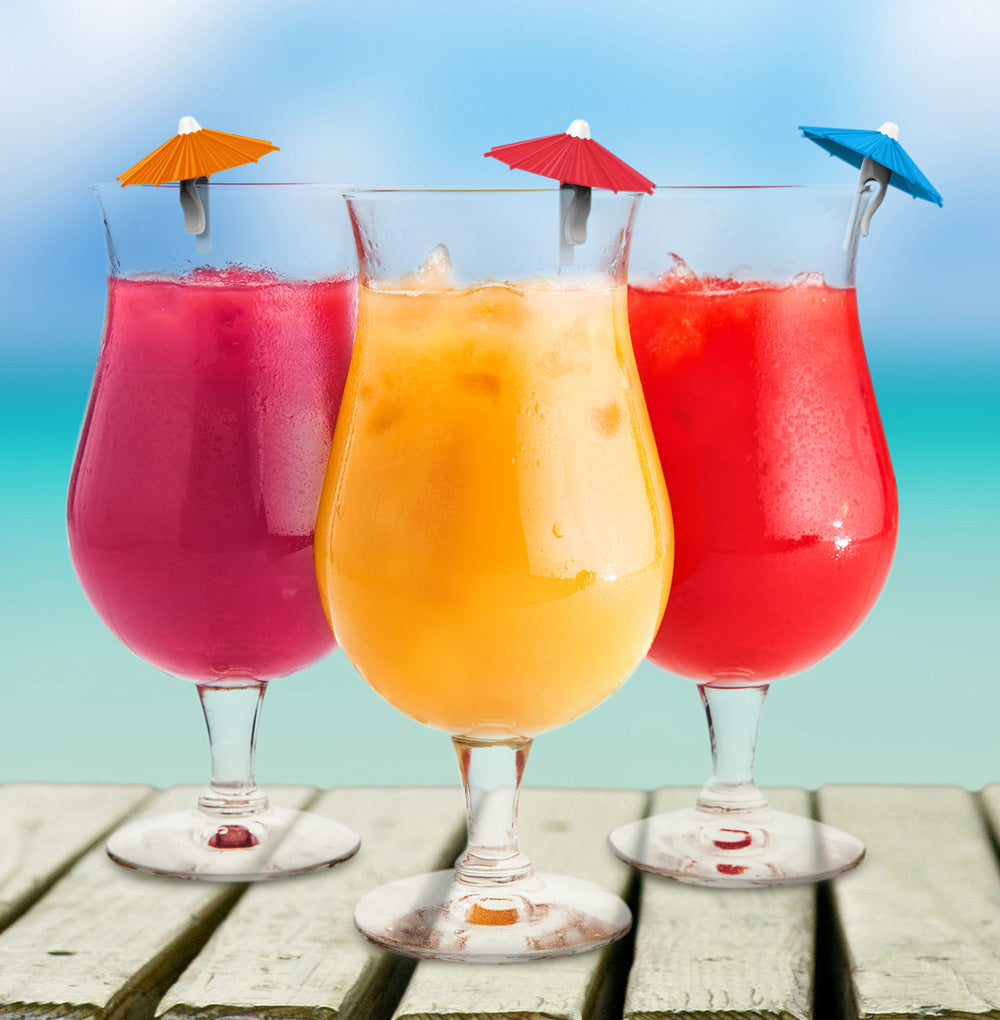 Three tall stemmed glasses filled with colorful tropical drinks, garnished with small paper umbrellas, set on a wooden surface against a blurred beach background.