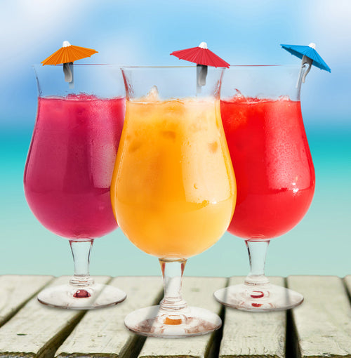 Three tall stemmed glasses filled with colorful tropical drinks, garnished with small paper umbrellas, set on a wooden surface against a blurred beach background.