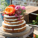 Multi-tiered cake with white frosting and colorful flowers on a wooden table outdoors.