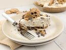 A slice of chocolate chip cookie pie on a beige speckled plate with a fork, topped with cookie dough and chocolate chips, with the whole pie blurred in the background on a white wooden table.