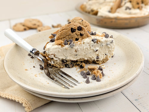 A slice of chocolate chip cookie pie on a beige speckled plate with a fork, topped with cookie dough and chocolate chips, with the whole pie blurred in the background on a white wooden table.