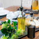 Two elegant glass bottles with pourer stoppers, featuring textured rings and an embossed logo, one half-filled with yellow liquid, possibly oil, and the other with a darker liquid, placed on a table with a cloth and some greenery in the background.