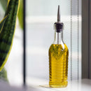 A glass bottle with a pourer stopper filled with a golden liquid - possibly oil - on a white surface with a plant nearby.