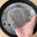 Hand holding a metal chain scrubber over a pot with soapy water.