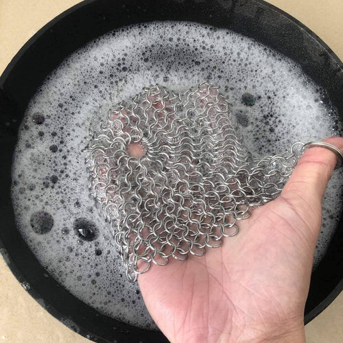 Hand holding a metal chain scrubber over a pot with soapy water.