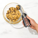 Cookie dough with chocolate chips in a glass bowl and a hand holding a metal cookie scoop.