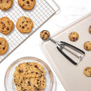 Chocolate chip cookies on a cooling rack, cookie dough on a plate, and baking sheet with cookie scoop.