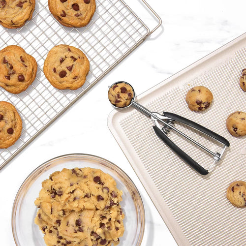 Chocolate chip cookies on a cooling rack, cookie dough on a plate, and baking sheet with cookie scoop.