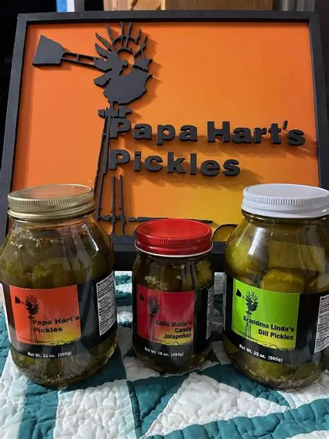 Three glass jars of Papa Hart's Pickles lined up in front of a branded orange sign with a black silhouette logo. The jars contain green pickled vegetables and have gold, red, and white lids, with colorful labels.
