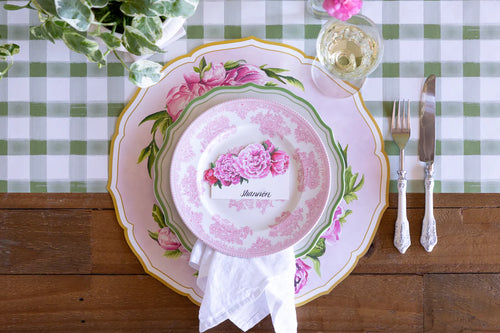 A green plaid table runner on a wooden surface with a place setting featuring peony placemat, plate, wine glass, silverware, and peony place card.