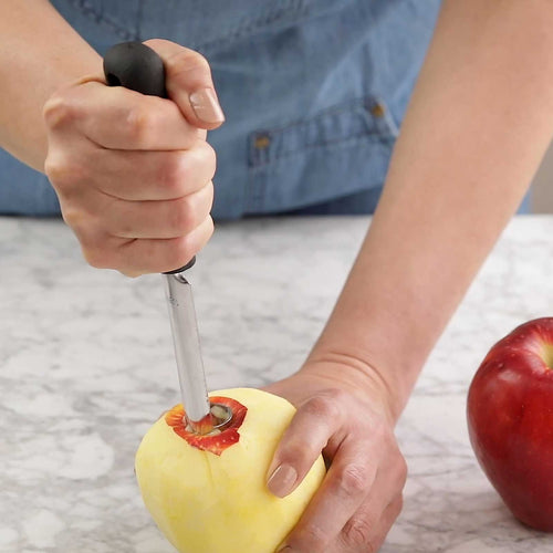 Person using an apple corer on a peeled apple with a red apple in the background.