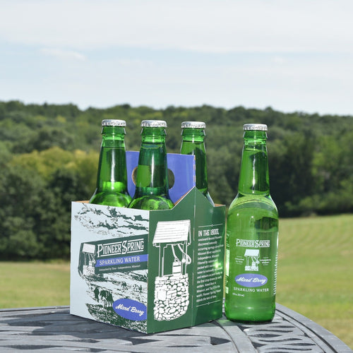 A case of Pioneer Spring Sparkling Water glass bottles placed on a table with a field in the background.