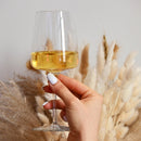 A person holding a wine glass filled with amber liquid with feathers and a neutral wall in the background.