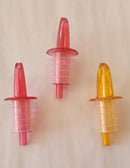 Three plastic bottle pourers in red, clear, and yellow on a beige background