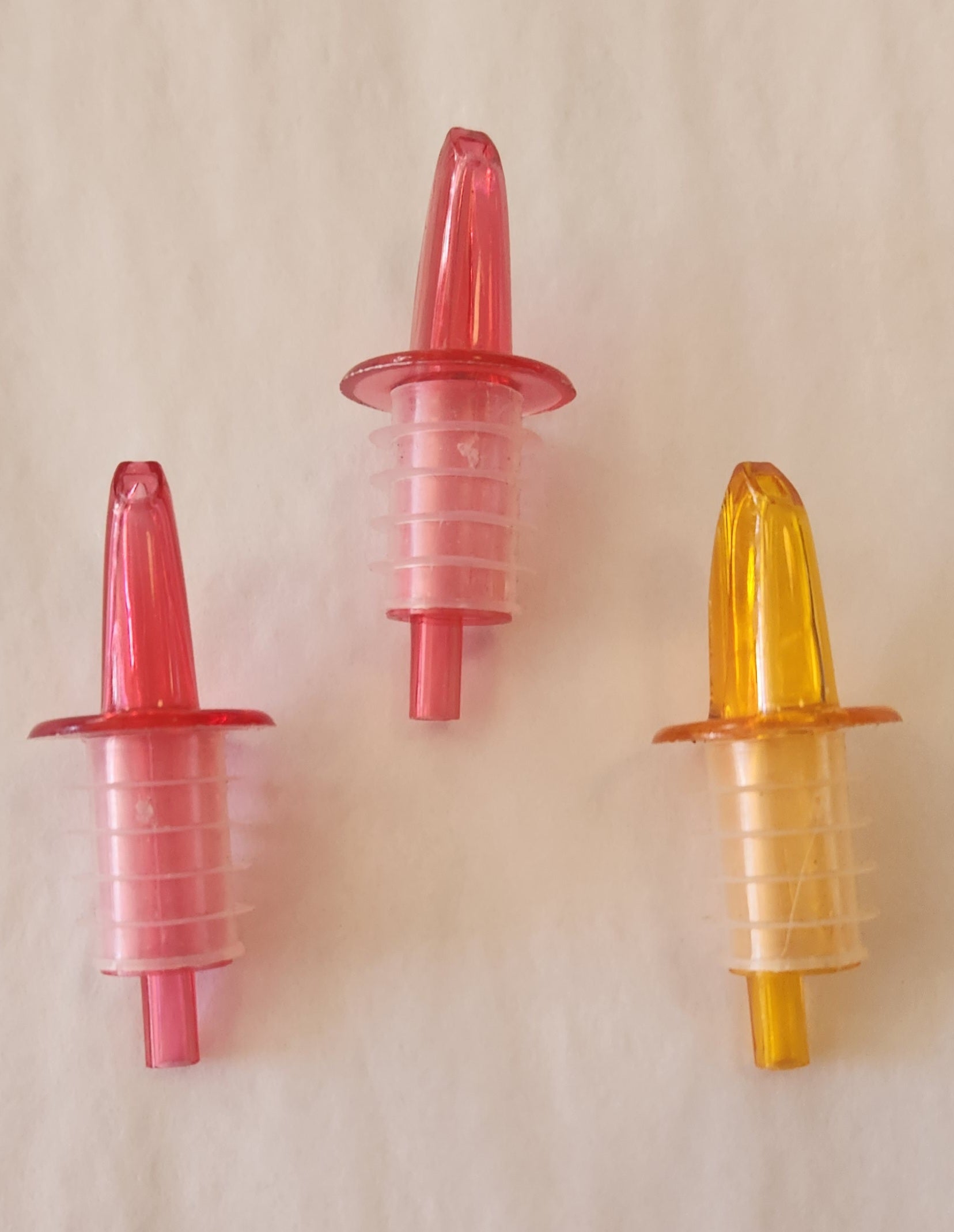 Three plastic bottle pourers in red, clear, and yellow on a beige background