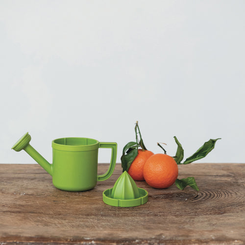 A bright green plastic watering can and matching citrus juicer set arranged on a rustic wooden table, with two oranges and green leaves beside them, against a plain light gray background.