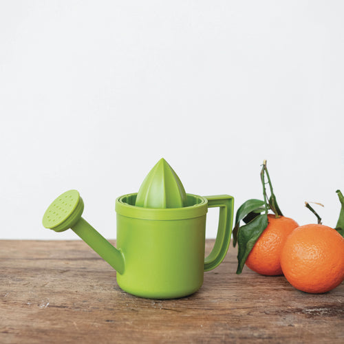 A bright green manual citrus juicer designed in the shape of a watering can, shown on a rustic wooden table next to fresh oranges with green leaves, against a clean white background.
