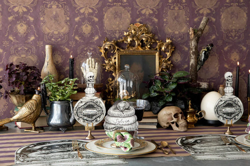 A gothic-themed tabletop display featuring paper skull-topped perfume bottle table decorations, a skeleton hand, a skull, candles, plants, and ornate gold wallpaper in the background.