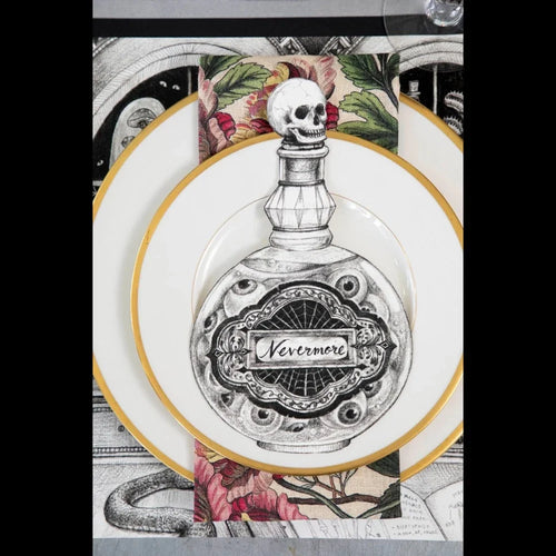 A decorative dinner plate featuring a black and white illustration of an ornate perfume bottle with a skull on the stopper and the word 'Nevermore' on the label. The plate is layered on top of a gold-rimmed plate and set against a patterned placemat with gothic illustrations.
