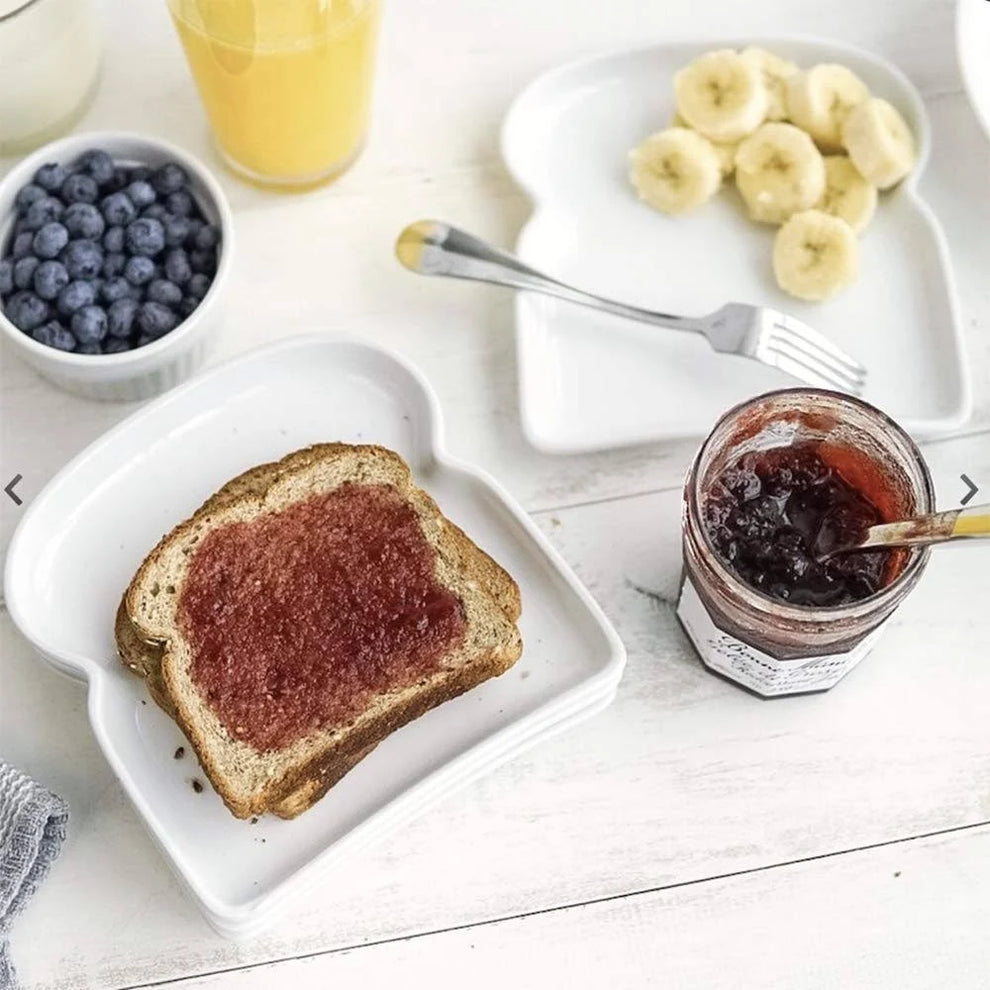 Plates shaped like pieces of toast with toast and bananas on them with a bowl of blueberries, jar of jam, and juice on a white wooden surface.