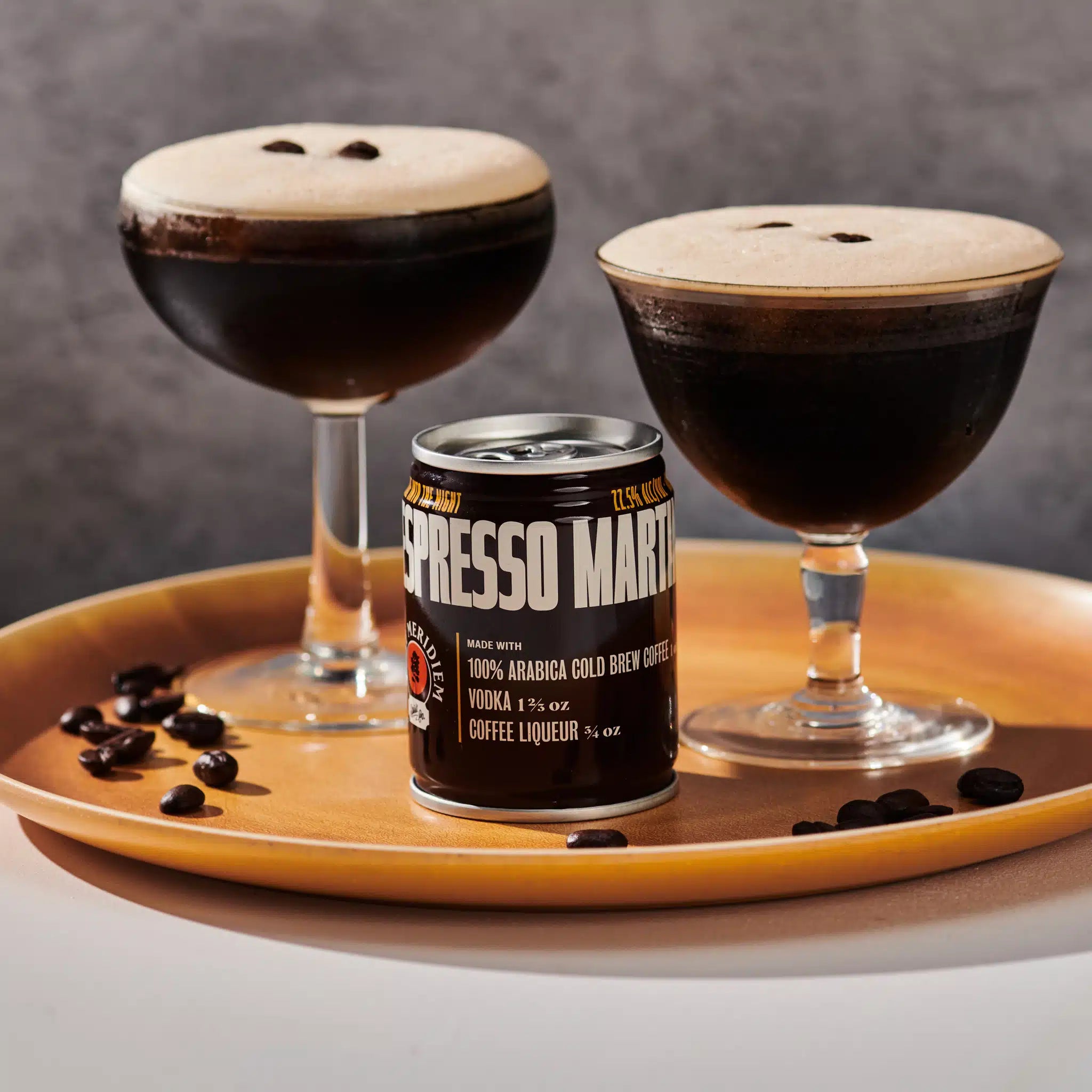A can of Espresso Martini coffee liqueur sits on a round wooden tray beside two dark espresso martinis in coupe glasses, with scattered coffee beans on a gray studio background.