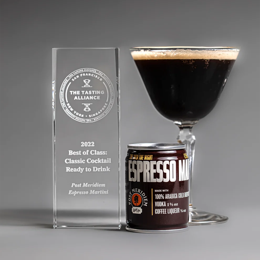 A glass award crystal plaque, a can of Espresso Martini liqueur, and a prepared espresso martini in a coupe glass, arranged on a gray studio background.