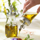 A person pouring oil from a bottle with a metal pour spout with a second bottle sitting on a white surface.