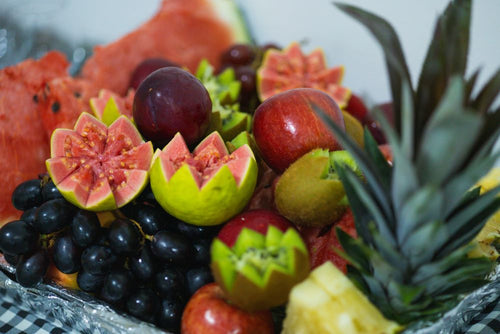 A close-up of a fruit assortment arranged in a bowl, including sliced watermelon, guava, grapes, apples, kiwi, and a pineapple, with a blurred light background.