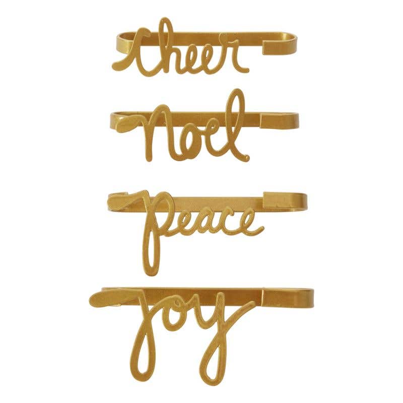A set of four gold-colored napkin holders with the words 'Cheer', 'Noel', 'Peace', and 'Joy' inscribed on them.