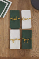 Four golden napkin holders with cursive words; 'noel, cheer, peace, joy' around green and white napkins on a wooden surface.