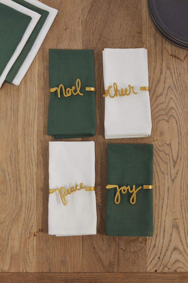 Four golden napkin holders with cursive words; 'noel, cheer, peace, joy' around green and white napkins on a wooden surface.