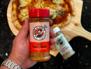 Person holding a bottle of Red Pizza Mojo seasoning above another bottle of seasoning and a pizza on a wood pizza peel.