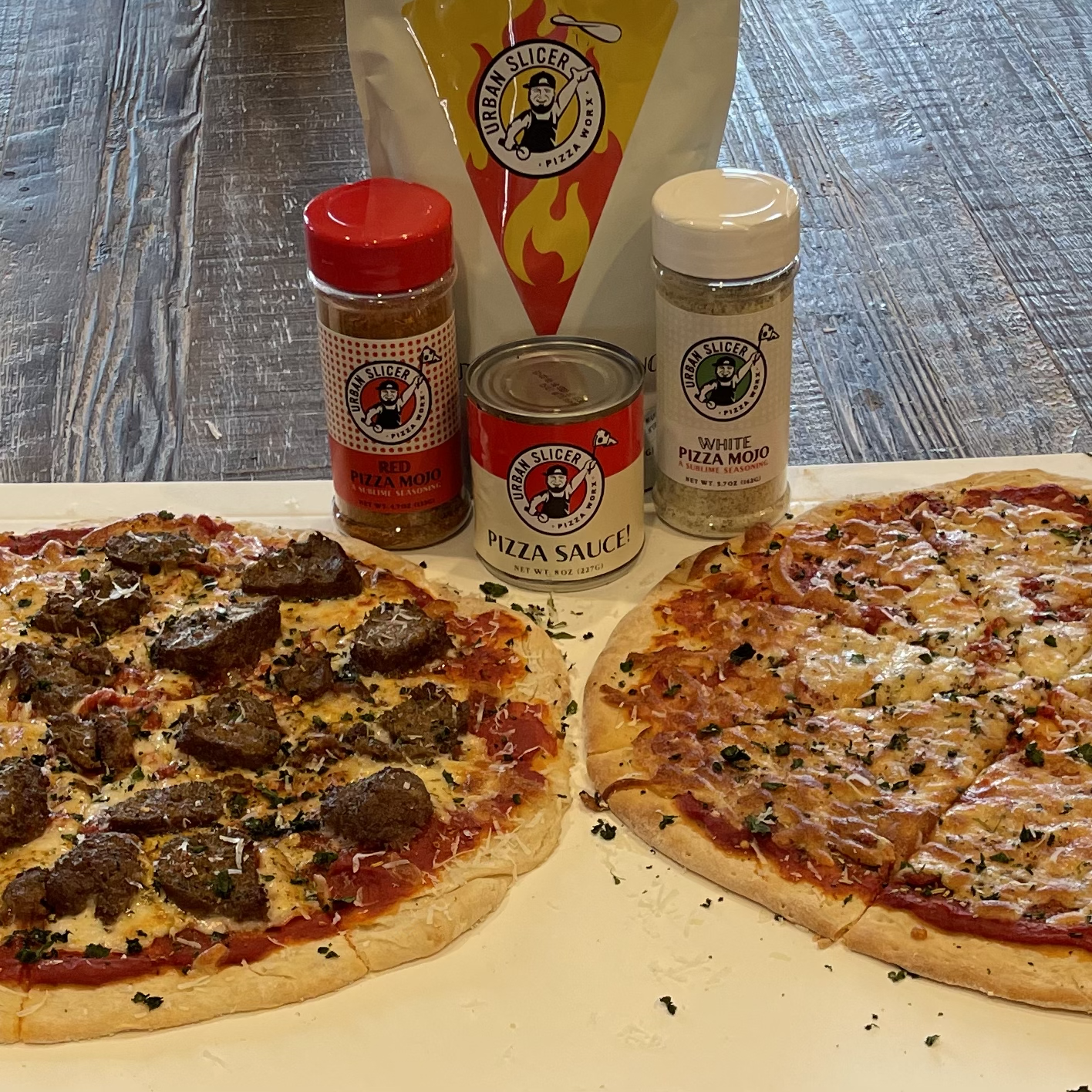 Two pizzas on a neutral surface with pizza seasonings and sauce next to them all on a wooden table.