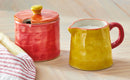 Red ceramic container with a yellow lid and a yellow pitcher on a wooden surface.