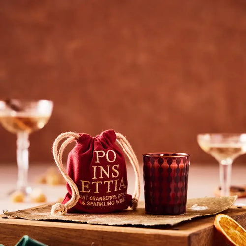 A small poinsettia scented candle next to a small drawstring bag with 'POINSETTIA' stitched on it, both on a wooden block with blurry champagne glasses with a blurred orange background.