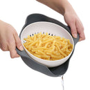 Person holding a white colander with gray handles filled with cooked pasta on a white background